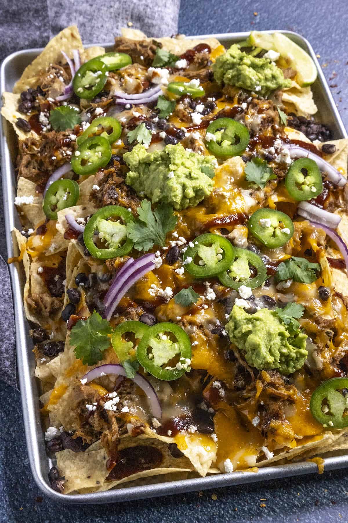 Serving up pulled pork nachos on a large tray, loaded with toppings, including guacamole, sliced jalapenos, sliced red onion, and bbq sauce