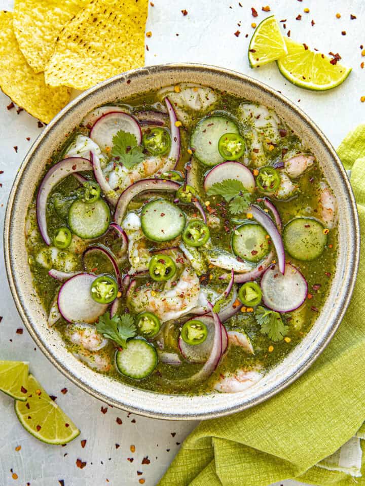 A bowl of aguachile features shrimp, cucumber slices, red onions, and cilantro in a vibrant green sauce. Lime slices and tortilla chips accompany this delicious aguachile recipe on a light surface with a green cloth.