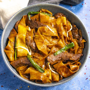A bowl of wide, flat noodles mixed with slices of beef, green onions, and bean sprouts offers a delicious beef chow fun experience. The dish is coated in a rich, dark sauce.