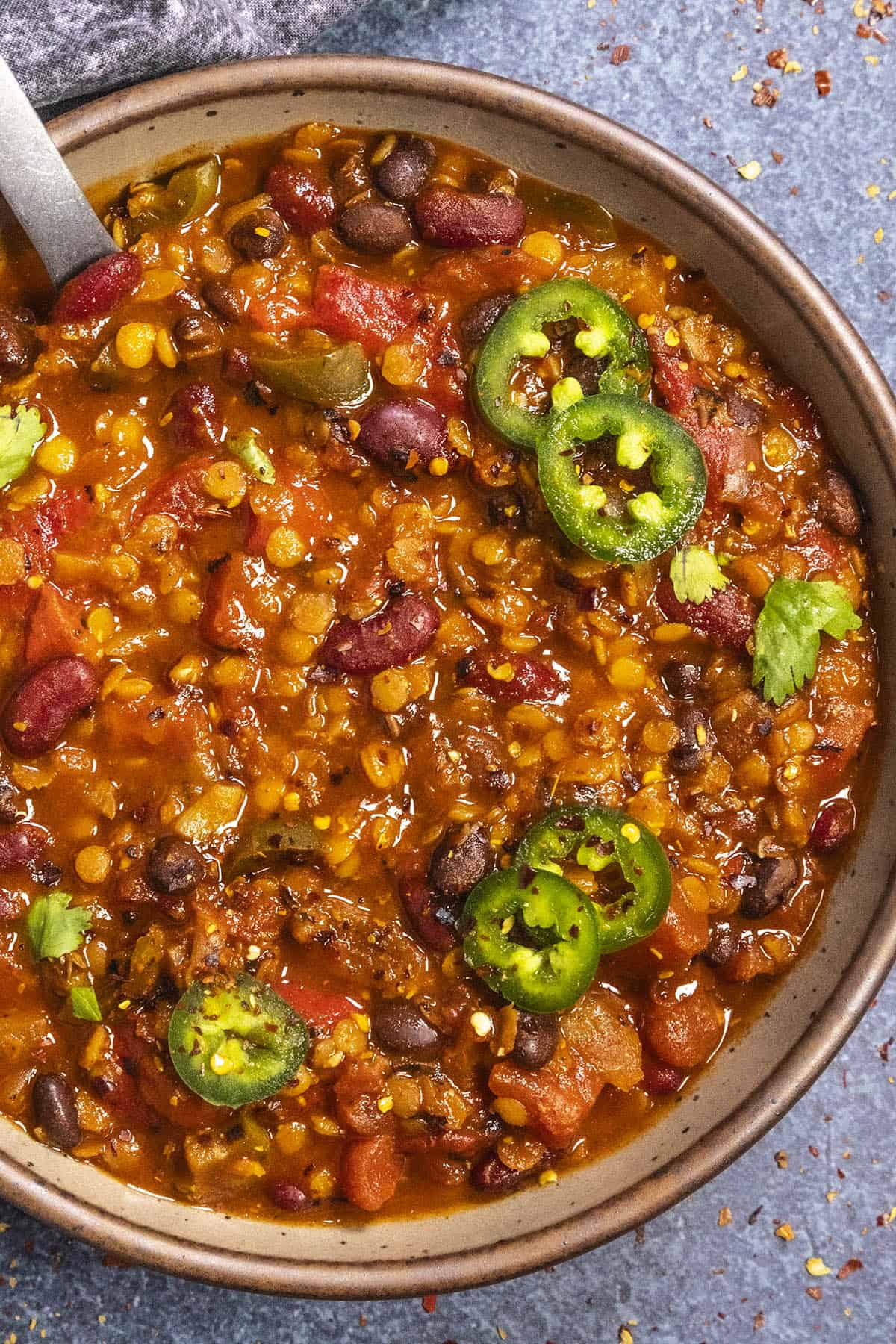 A bowl of lentil chili filled with lentils, beans and tomatoes, garnished with sliced jalape&ntilde;os and cilantro. A spoon rests inside the bowl, which is placed on a textured surface with a cloth nearby.