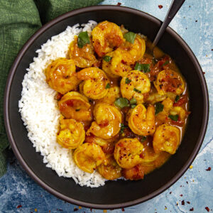 A bowl of white rice topped with shrimp in a yellow, spiced sauce with chopped green herbs and red pepper flakes, served on a blue surface with a green napkin nearby.