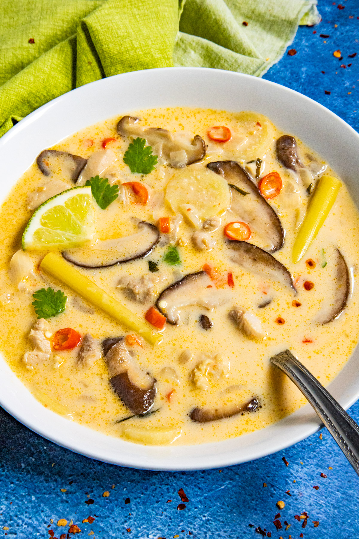 A bowl of creamy Thai coconut chicken soup, or tom kha gai, with sliced mushrooms, chili peppers, lemongrass, a lime wedge, and cilantro sits on a blue surface. A spoon rests in the bowl while a green cloth adds contrast in the background.