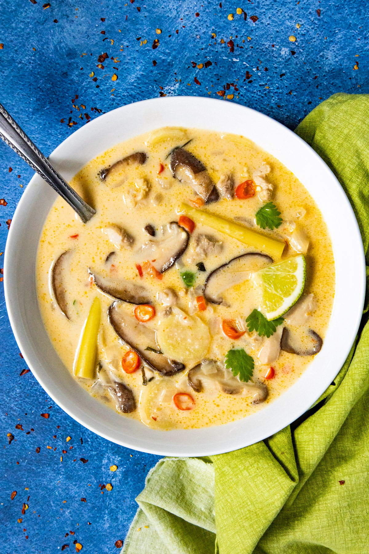A bowl of creamy Tom Kha Gai soup with sliced mushrooms, herbs, and red chili slices on a blue surface. A lime wedge and a green cloth are beside the bowl.