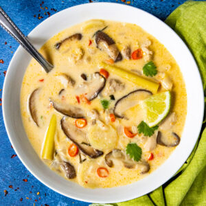 A bowl of creamy tom kha gai garnished with sliced red chili, lime wedge, and fresh cilantro on a blue surface. A spoon is placed in the bowl, and a green napkin is beside it.