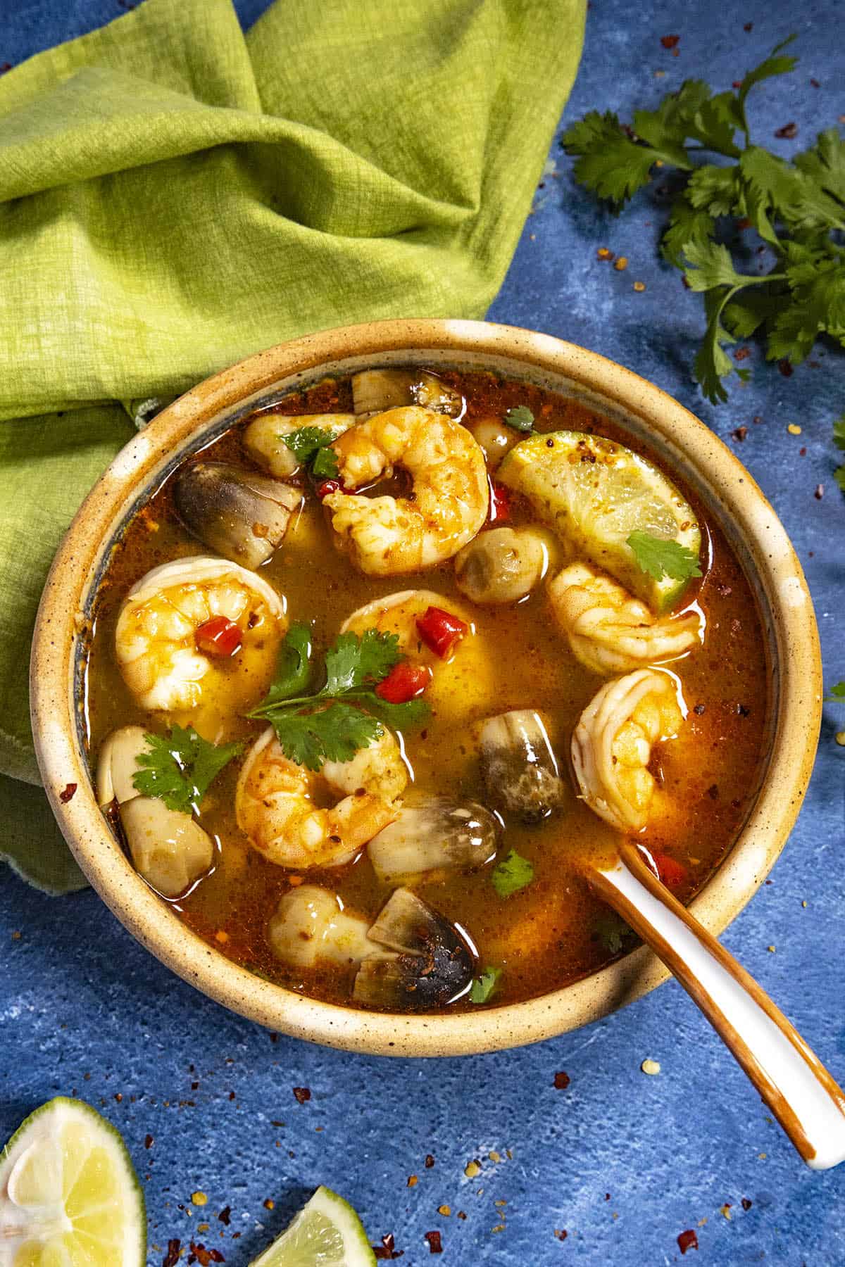 A large bowl of spicy Tom Yum Soup loaded up with shrimp and mushrooms, garnished with spicy red chilies, cilantro, and lime