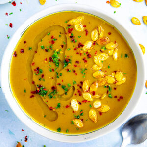 A bowl filled with spicy and creamy pumpkin soup, topped with roasted pumpkin seeds, chili flakes, and a drizzle of oil