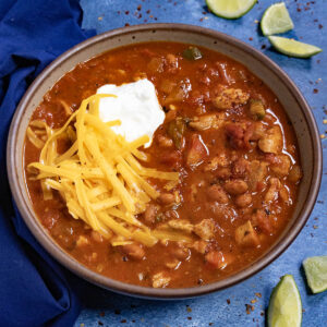 A large bowl of chicken chili, topped with freshly shredded cheddar cheese and sour cream, with lime wedges on the side